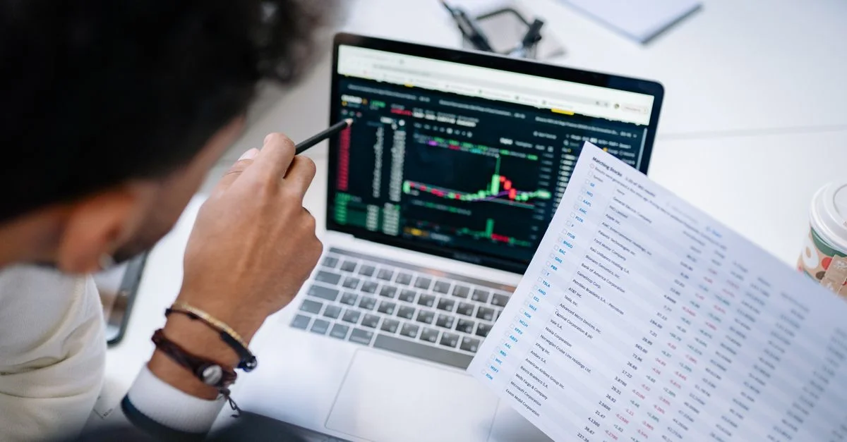 Un professionnel analyse des données boursières sur l'écran d'un ordinateur portable avec des documents financiers.